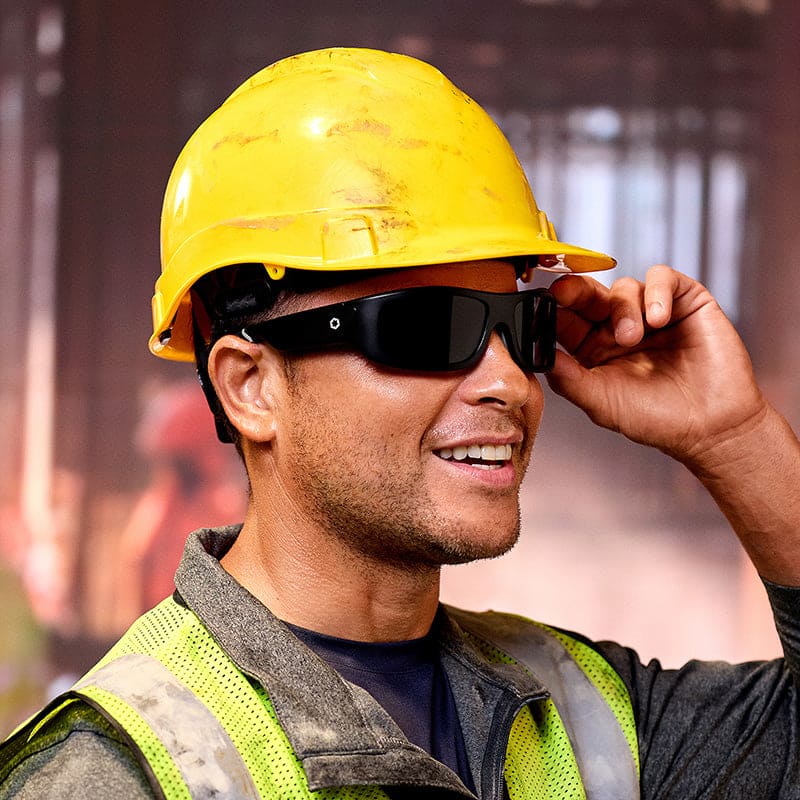 A smiling construction worker in a yellow hard hat and reflective vest adjusts his Lucyd Armor | Black Polarized sunglasses, which feature ANSI Z87.1-certified tactical-grade protection. The background is blurred.