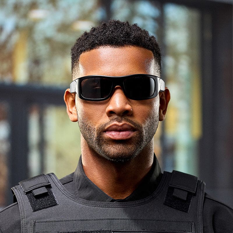 A man wearing Lucyd Armor | Black Polarized sunglasses and a black tactical vest stands outdoors with a serious expression, looking at the camera. The background is blurred with hints of trees and a building.