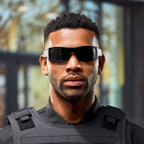 A man wearing Lucyd Armor | Black Polarized sunglasses and a black tactical vest stands outdoors with a serious expression, looking at the camera. The background is blurred with hints of trees and a building.