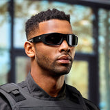 A man with short curly hair and a trimmed beard wears Lucyd Armor | Black Polarized sunglasses by Lucyd and a black tactical vest, standing indoors in front of blurred windows and trees.