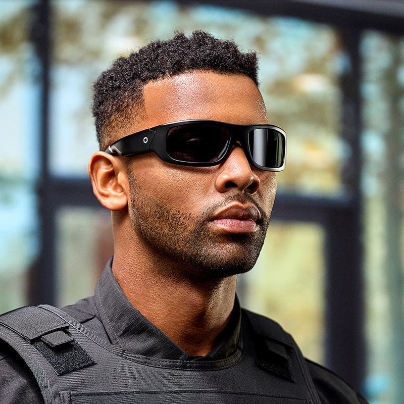 A man with short curly hair and a trimmed beard wears Lucyd Armor | Black Polarized sunglasses by Lucyd and a black tactical vest, standing indoors in front of blurred windows and trees.