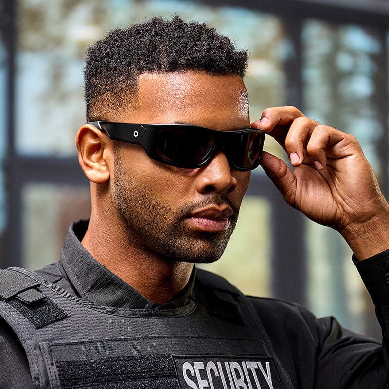A man in a black security uniform stands indoors, adjusting his Lucyd Armor | Black Polarized sunglasses by Lucyd. The softly blurred background features light streaming through large windows.