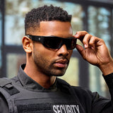 A man in a black security uniform stands indoors, adjusting his Lucyd Armor | Black Polarized sunglasses by Lucyd. The softly blurred background features light streaming through large windows.