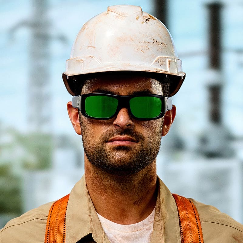 A construction worker wearing a white hard hat, tan shirt, orange suspenders, and Lucyd Armor | Green Mirror Polarized sunglasses by Lucyd stands outdoors with a blurred industrial background.