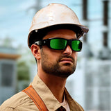 A construction worker wearing Lucyd Armor | Green Mirror Polarized safety glasses by Lucyd, a white hard hat, and a tan shirt stands outdoors with a serious expression; industrial equipment is blurred in the background.