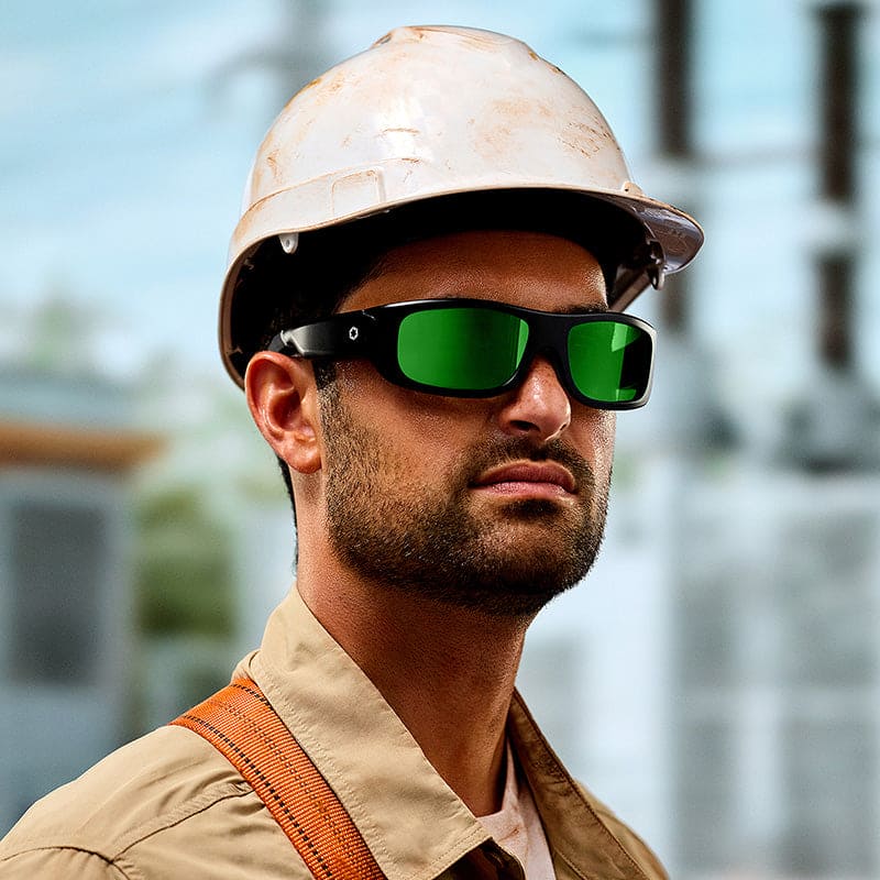 A construction worker wearing Lucyd Armor | Green Mirror Polarized safety glasses by Lucyd, a white hard hat, and a tan shirt stands outdoors with a serious expression; industrial equipment is blurred in the background.