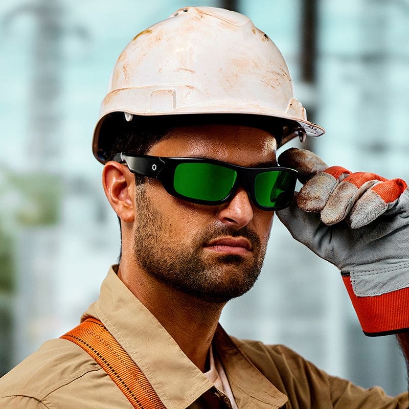 A construction worker wearing a white hard hat, gloves, a brown work shirt, and Lucyd Armor | Green Mirror Polarized safety glasses by Lucyd looks forward while adjusting his glasses. The background is blurred.
