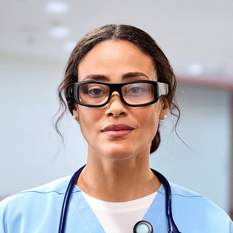 A woman in medical scrubs wears Lucyd Armor | Slim Photochromic eyewear by Lucyd and a stethoscope, looking confidently at the camera in a bright indoor setting.