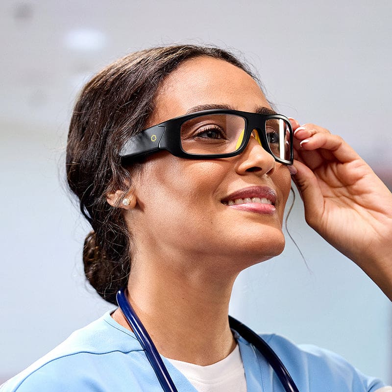 A woman in light blue scrubs and a stethoscope smiles as she adjusts Lucyd Armor | Slim Photochromic audio glasses by Lucyd in a brightly lit indoor setting.