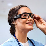 A woman in light blue scrubs and a stethoscope smiles as she adjusts Lucyd Armor | Slim Photochromic audio glasses by Lucyd in a brightly lit indoor setting.