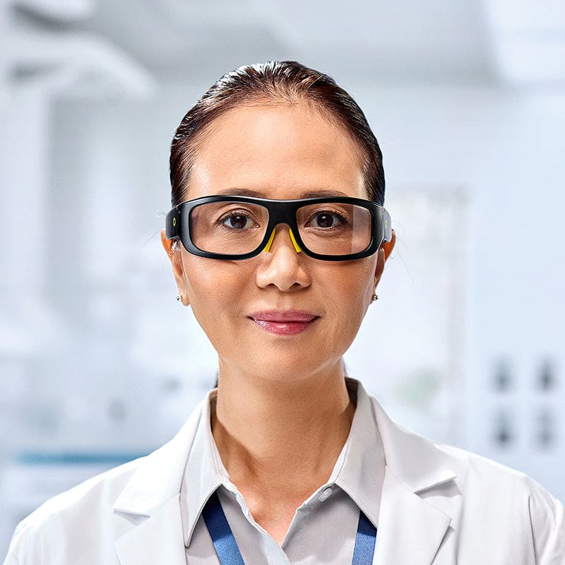 A woman wearing Lucyd Armor | Slim Photochromic safety glasses by Lucyd and a white lab coat stands in a bright lab, looking at the camera with a slight smile.