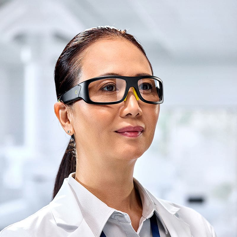 A woman in a white lab coat wears Lucyd Armor | Slim Photochromic glasses by Lucyd, standing confidently in a bright, modern laboratory.