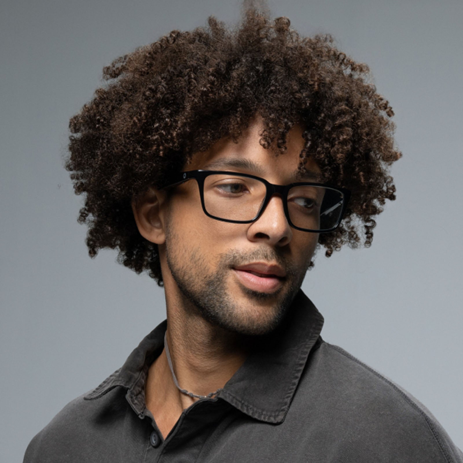 A man with curly dark hair, wearing Lucyd Darkside black rectangular Bluetooth audio sunglasses and a dark collared shirt, looks to the side against a plain gray background.