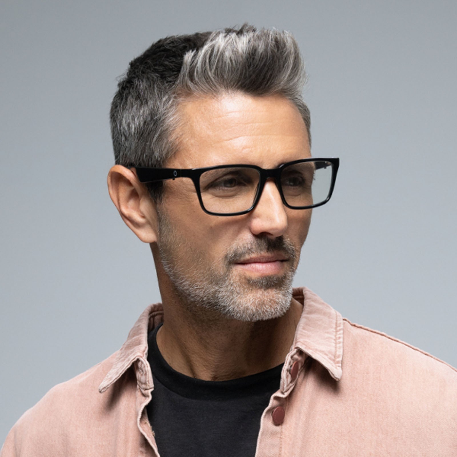 A man with short salt-and-pepper hair and a trimmed beard wears Lucyd Darkside black Bluetooth audio sunglasses, a black shirt, and a light pink jacket, looking to the side against a plain gray background.