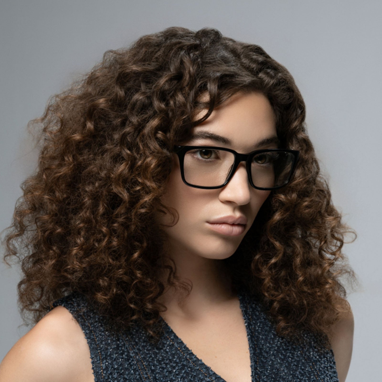 A woman with thick, curly brown hair wears Lucyd Darkside large black Bluetooth audio sunglasses, looking slightly to the side in a textured dark sleeveless top against a neutral gray background.