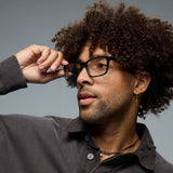 A person with curly hair, wearing Lucyd Darkside black Bluetooth audio sunglasses, a brown collared shirt, a cross earring, and a necklace adjusts their glasses while looking to the side against a gray background.