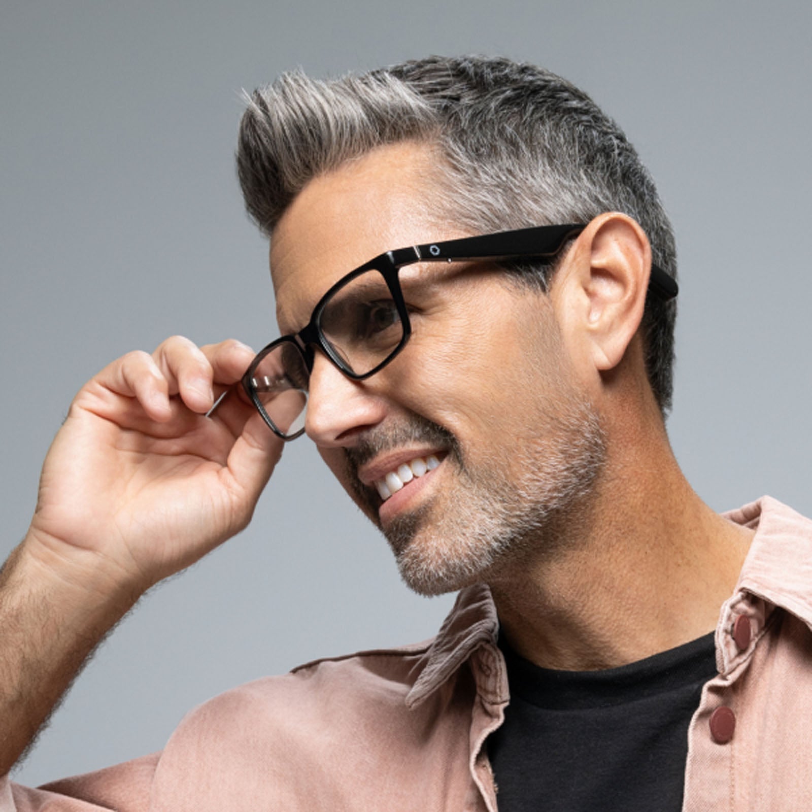 A smiling man with short gray hair and stubble adjusts his Lucyd Darkside Bluetooth audio sunglasses. He wears a light pink button-up shirt over a black t-shirt, set against a plain light gray background.