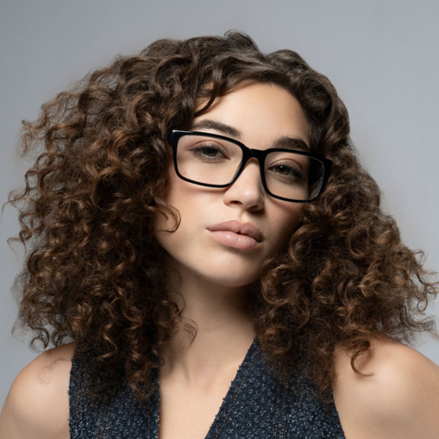 A young woman with curly brown hair wears Lucyd’s Darkside black rectangular Bluetooth audio sunglasses, looking at the camera with a neutral expression. She pairs them with a sleeveless textured top against a plain gray background.