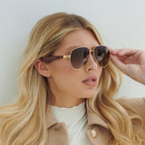 A woman with long blonde hair wears Lucyd XL Jupiter XXL brown Bluetooth audio sunglasses with polarized lenses and a beige coat over a white top, posing against a light-colored background.