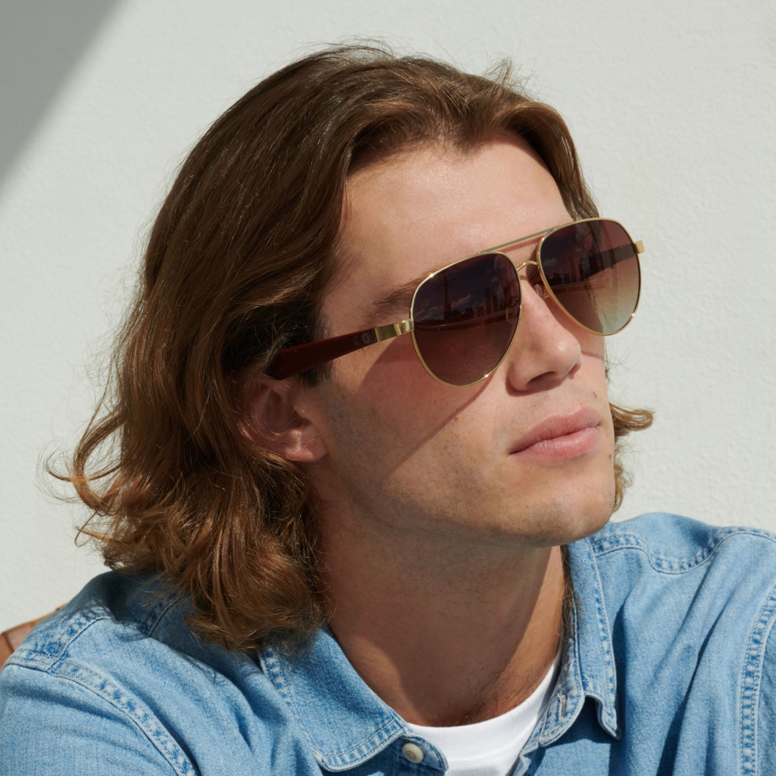 A young man with shoulder-length brown hair and a light denim shirt sits against a white wall, wearing Lucyd XL Jupiter XXL Bluetooth audio sunglasses with polarized lenses, looking up with a relaxed expression.
