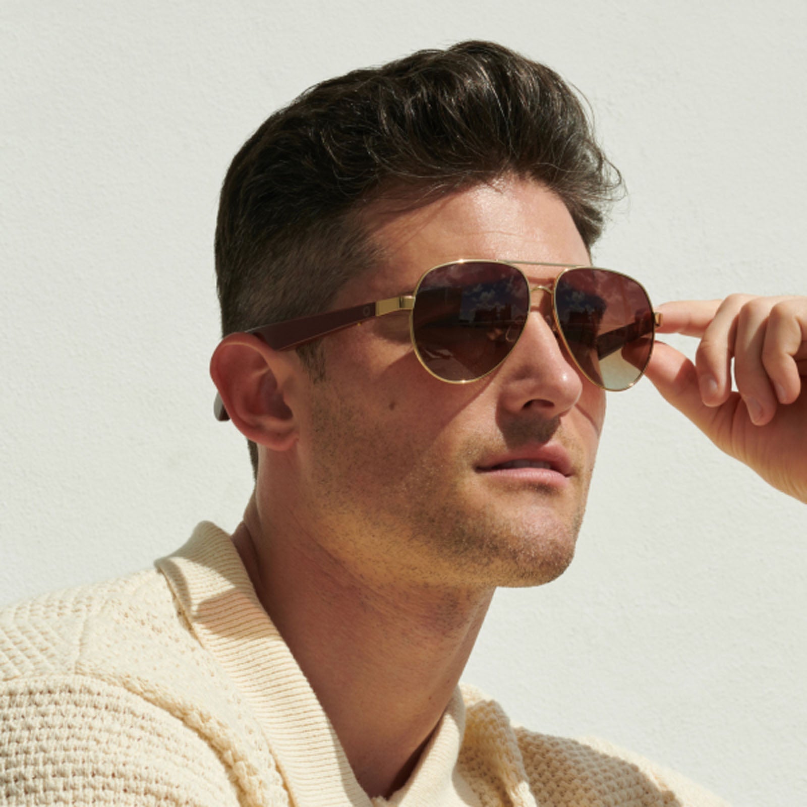 A man with short brown hair, wearing Lucyd XL Jupiter XXL Bluetooth audio sunglasses and a cream knit sweater, holds the frame of his sunglasses while looking off into the distance against a plain white background.