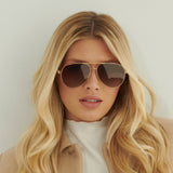 A confident woman with long blonde hair wears Lucyd XL Jupiter XXL Bluetooth audio sunglasses with polarized lenses, paired with a light turtleneck and tan coat, standing against a pale background and looking at the camera.