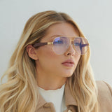 A young woman with long blonde hair wears Lucyd XL’s Jupiter XXL gold-rimmed polarized eyeglasses and a beige coat, gazing thoughtfully to the side against a plain light background.