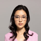 A woman with long dark wavy hair, wearing Lucyd Moonbeam AI smart glasses and a light pink V-neck sweater, looks directly at the camera against a plain light gray background.