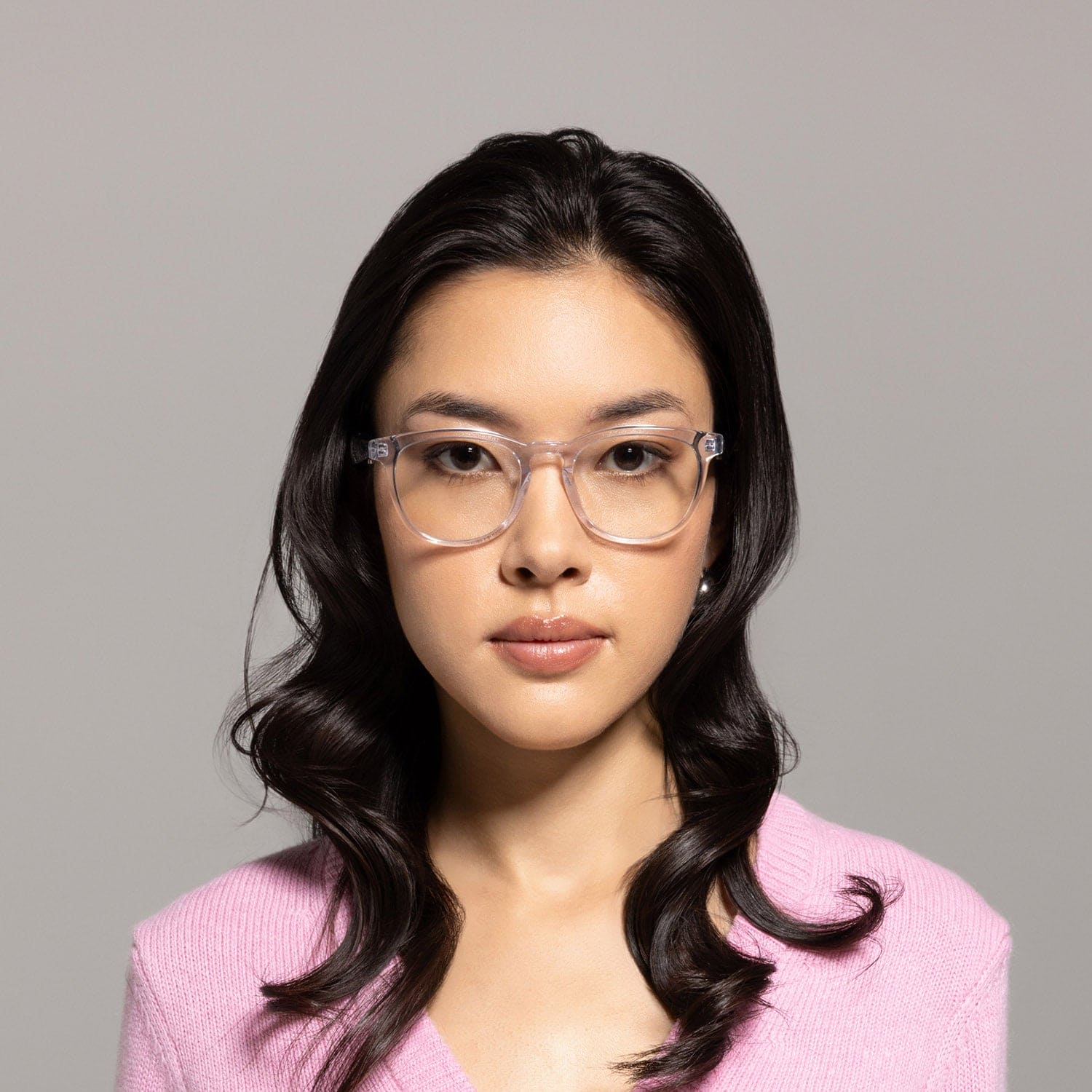 A woman with long dark wavy hair, wearing Lucyd Moonbeam AI smart glasses and a light pink V-neck sweater, looks directly at the camera against a plain light gray background.