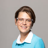 A smiling person with short brown hair wears Lucyd Moonbeam smart glasses with clear photochromic lenses, a blue sweater over a white collared shirt, and small earrings, posing against a plain gray background.
