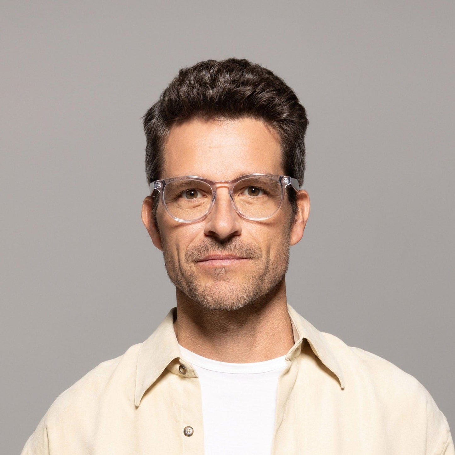 A man with short, dark hair and a neutral expression is wearing Lucyd's Moonbeam glasses with photochromic lenses, paired with a light beige shirt over a white t-shirt, against a plain gray background.