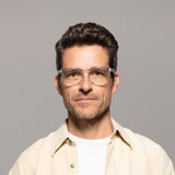 A man with short, dark hair and a neutral expression is wearing Lucyd's Moonbeam glasses with photochromic lenses, paired with a light beige shirt over a white t-shirt, against a plain gray background.