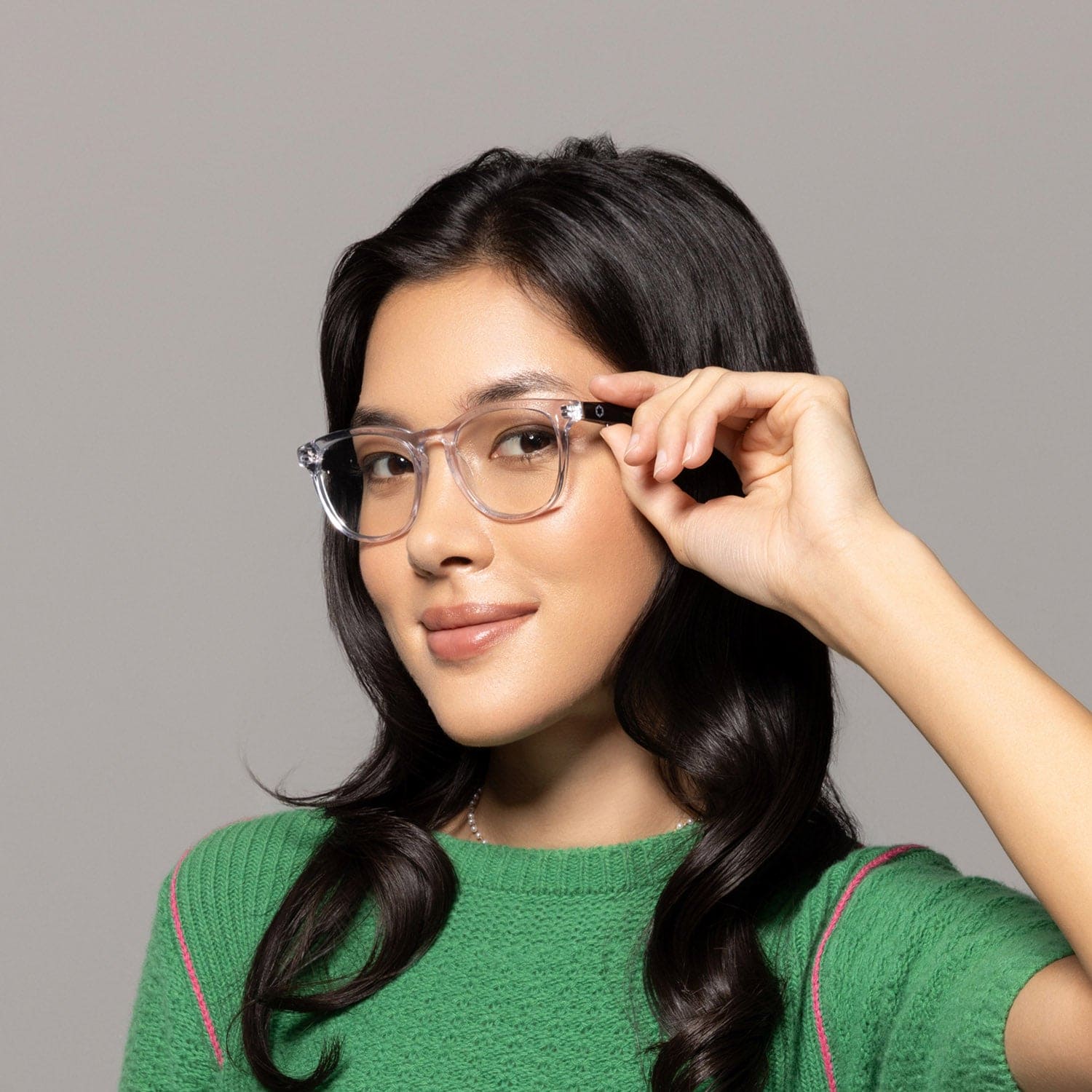 A woman with long dark hair, wearing Lucyd’s Moonbeam clear photochromic lenses and a green sweater, smiles softly while holding her glasses against a plain gray background.